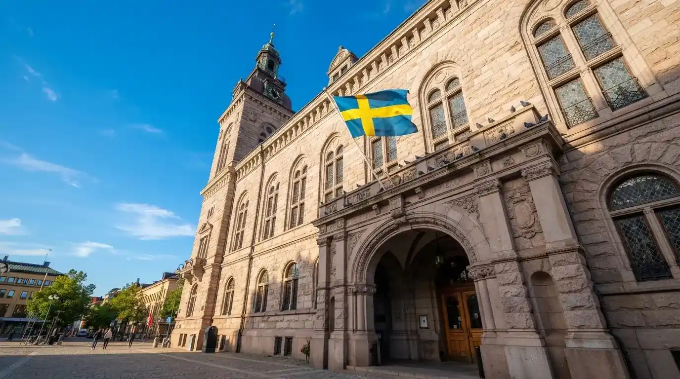 Ett gammalt stadshus i sten fotograferat underifrån mot en klarblå himmel, med den svenska flaggan som fladdrar ovanför entrén.